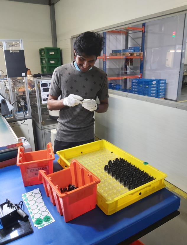 Operator in gloves inspecting small components from a tray at a line-side quality bench with bins and trays.