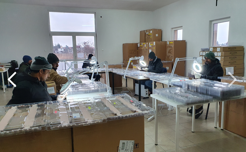 Several inspectors at long tables using magnifying lamps to examine clear molded plastic parts in a warehouse workspace.