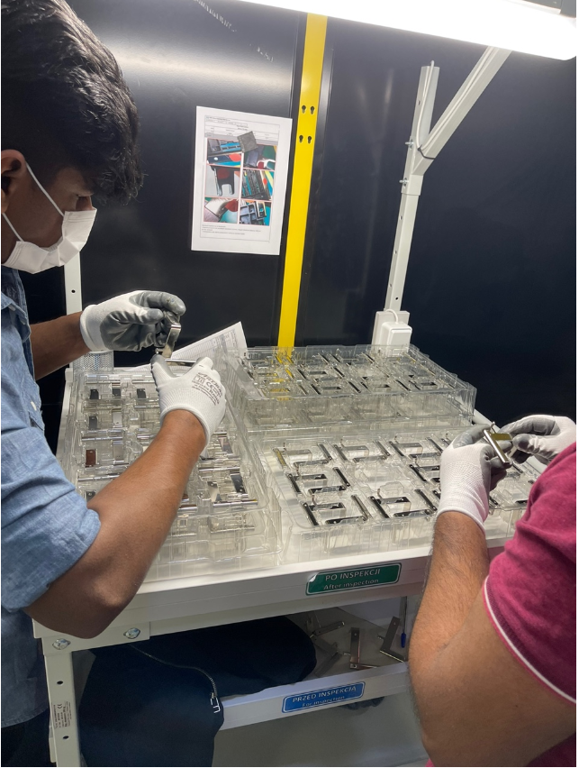 Two workers in masks and gloves sorting metal brackets at a workstation with before-and-after inspection shelf labels.