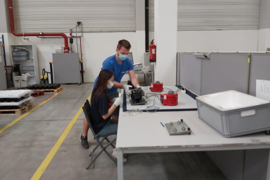 Two masked technicians inspecting a large mechanical assembly on a white bench with test equipment in a high-bay warehouse.