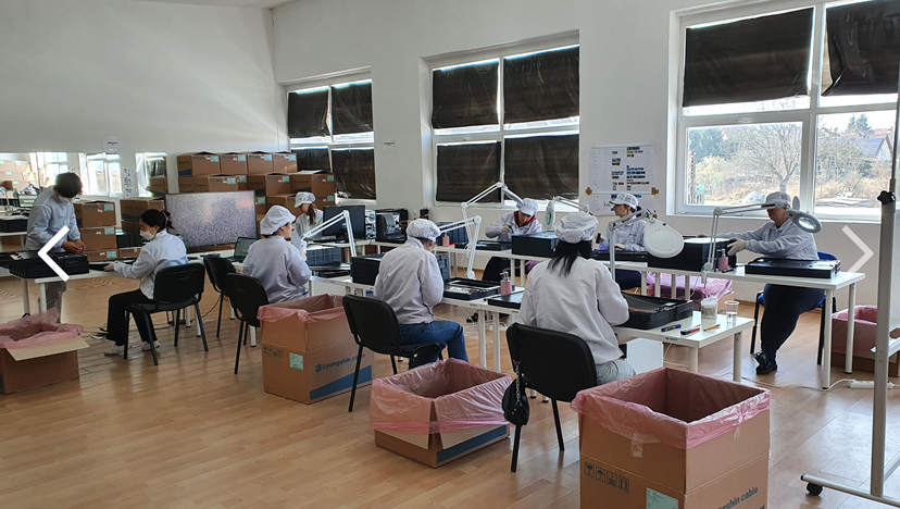 Team in lab coats and hairnets performing detailed inspection under magnifying lamps along a row of tables with shipping cartons.