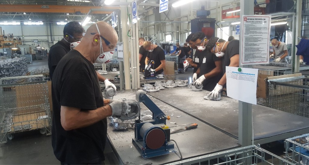 Line of operators in respirators and PPE performing manual finishing and inspection of die-cast metal parts at black workbenches.