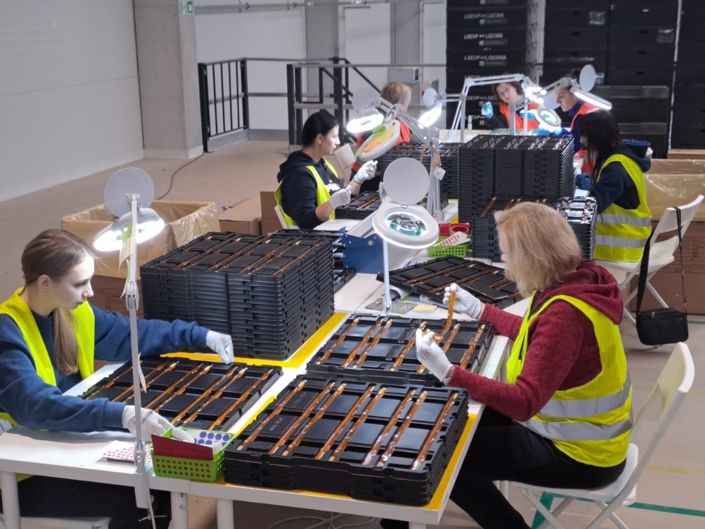 Inspectors in high-visibility vests examining trayed electronic-style parts under magnifying lamps at white tables.