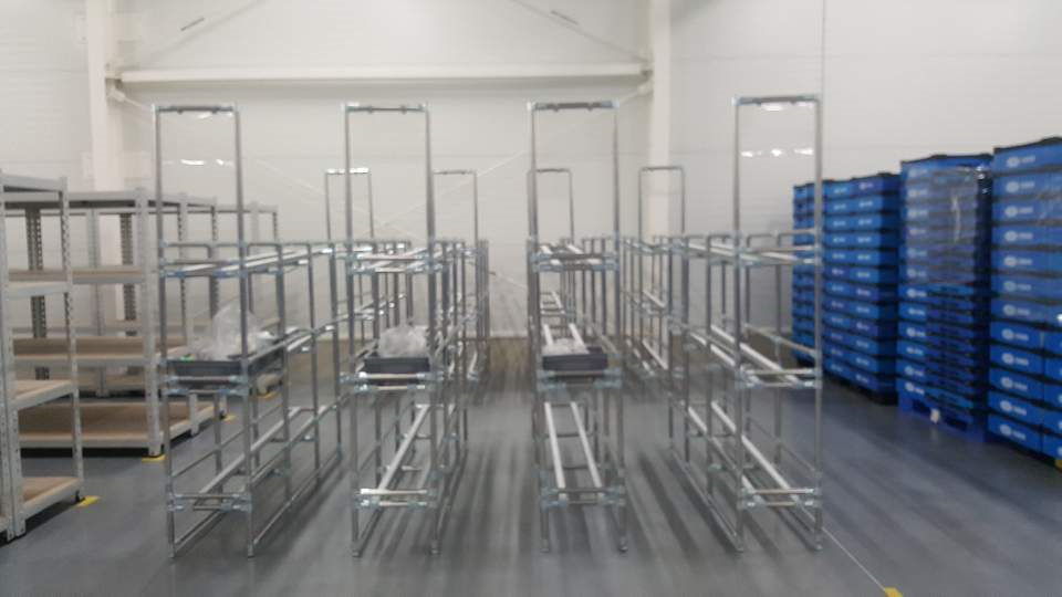 Silver pipe racks with roller tracks next to stacks of blue corrugated plastic boxes in a warehouse.