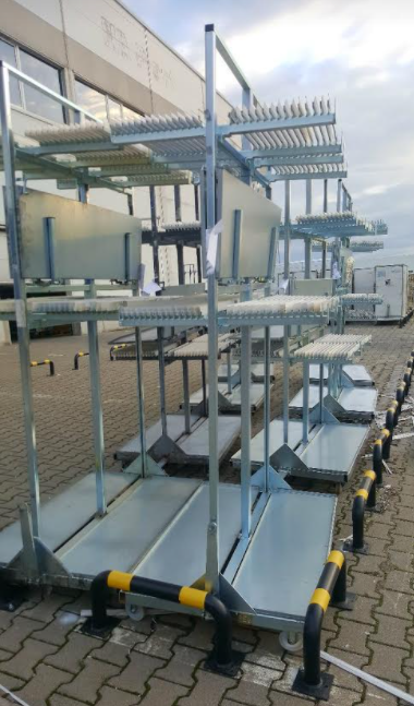 Tall galvanized mobile racks with horizontal arms and protective comb dividers staged outside a plant building.